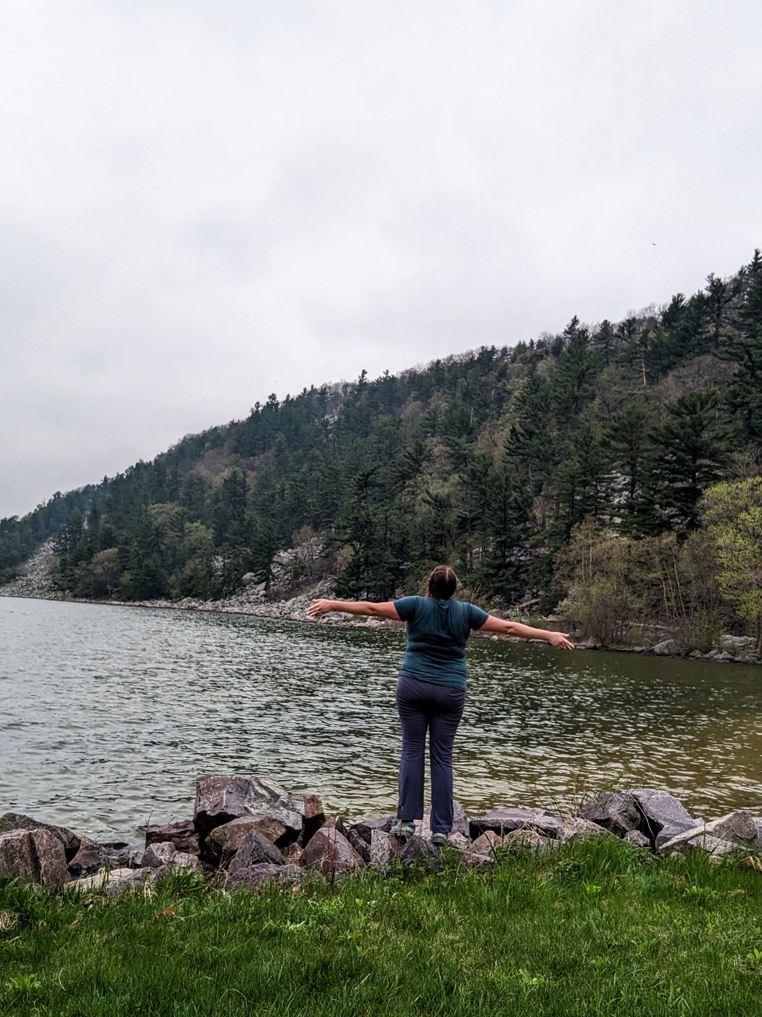 woman wearing hiking clothes stands with back to camera and arms outspread at the edge of a lake with a tree covered bluff in the background