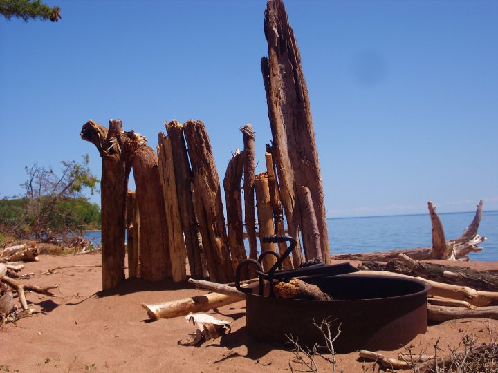 a firepit on the beach with a makeshift windbreak constructed from logs and driftwood in the sand