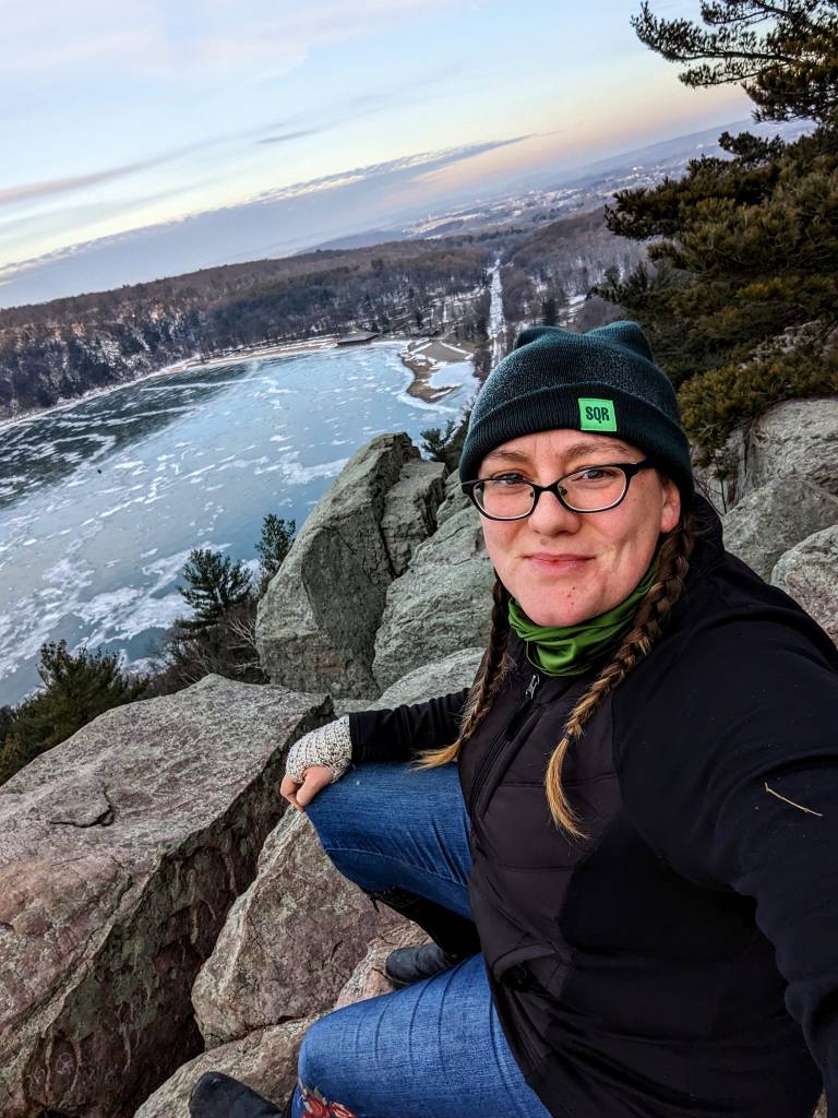 woman in winter coat and hat sits atop rocky outcropping looking at the camera with a frozen lake down below in the background
