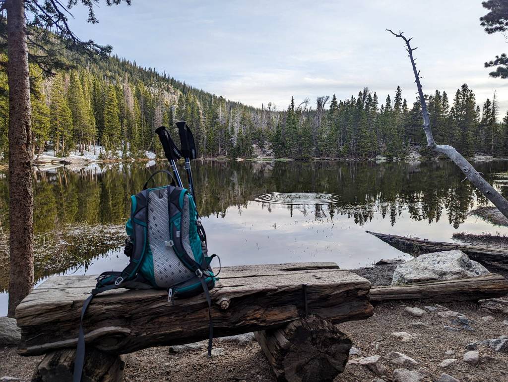 a backpack and trekking poles sits on a rustic log bench in front of an alpine lake