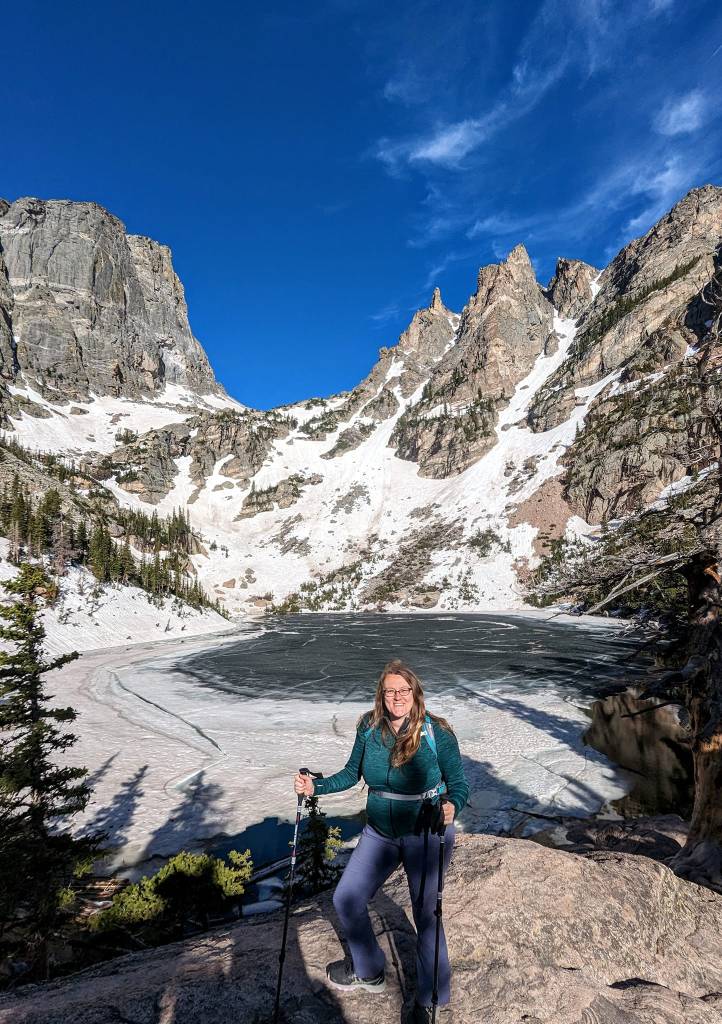 a woman in hiking clothes with a backpack and trekking poles stands in front of a frozen alpine lake surrounded by snowy mountain crags