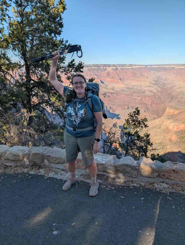 a female backpacker stands on the north rim of the Grand Canyon, triumphantly raising her hiking poles above her head